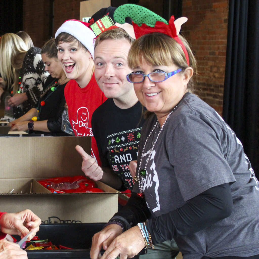 Three employees look up from working on a group service project and smile at the camera. The person in the middle is holding a thumbs up. They are all decked out in red and green holiday clothing and accessories.