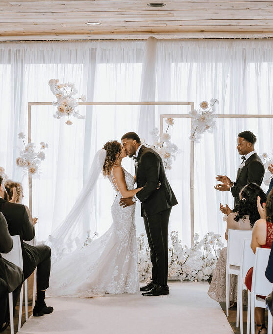 A couple stands at the end of their wedding aisle, kissing at the end of their ceremony. A best man is to the right of the groom and applauds as they embrace. Guests in chairs to the left and right applaud as well. Behind the couple are flowing white drapes with light coming through softly, and a white frame-like altar piece decorated with white flowers.