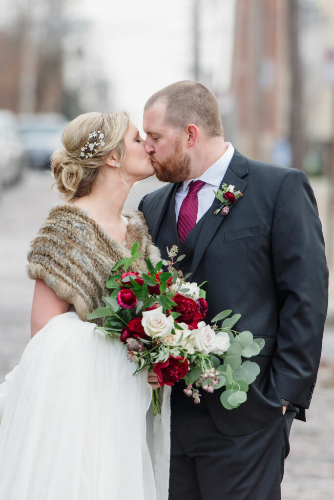 Chelsea and Chris share a kiss outside the High Line Car House in Columbus Ohio. Behind them is a blurred brick street and old urban buildings. Chelsea is wearing a brown fur shawl and carrying a bouquet of white, red, and green florals. Chris is in a black suit and red tie.