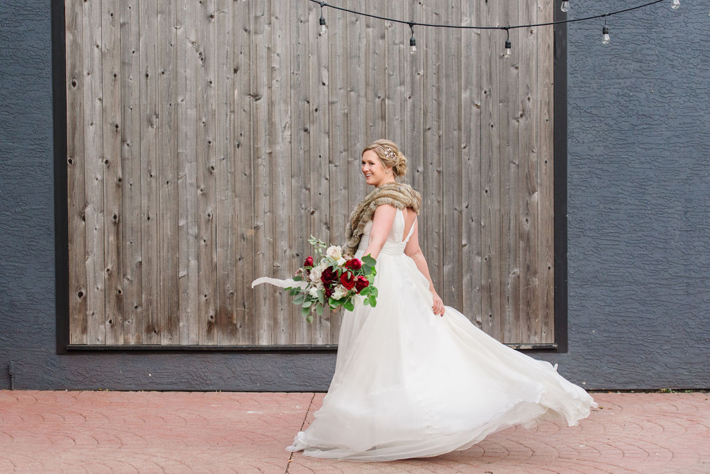 Chelsea is twirling outside the High Line Car House in her white wedding dress and brown fur shawl. She is smiling as she spins, her skirt in one hand and her bouquet of red, white, and green florals in the other. Behind her is a charcoal painted brick wall and weathered, industrial wood siding.