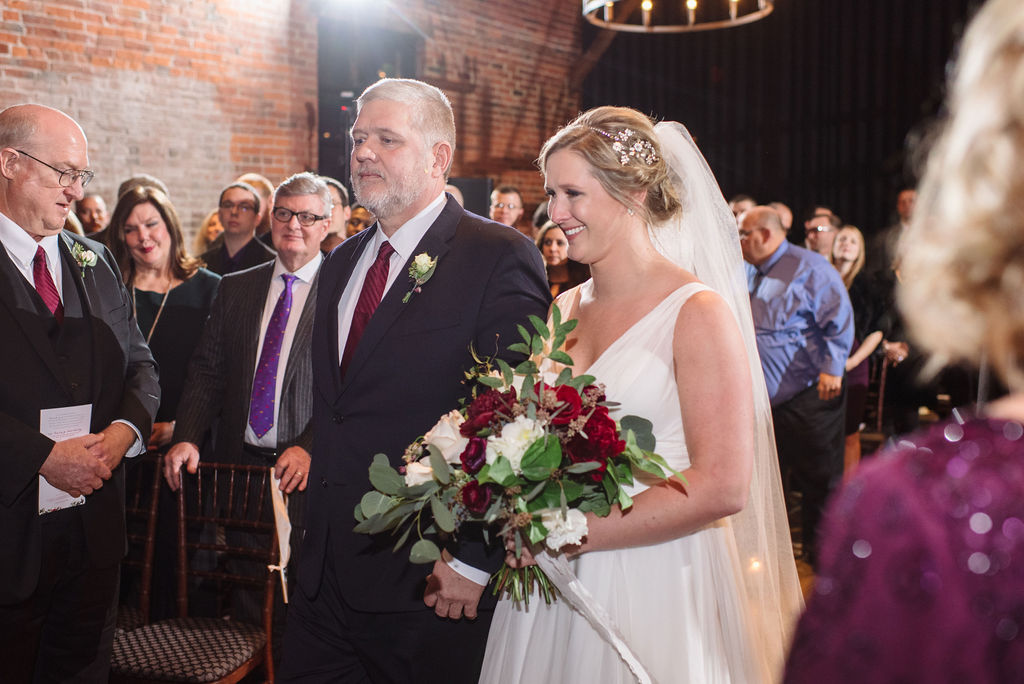 Chelsea is escorted down the aisle, smiling, with a bouquet of green, red, and white florals. Guests are all standing as she approaches the front of the room, out of frame.