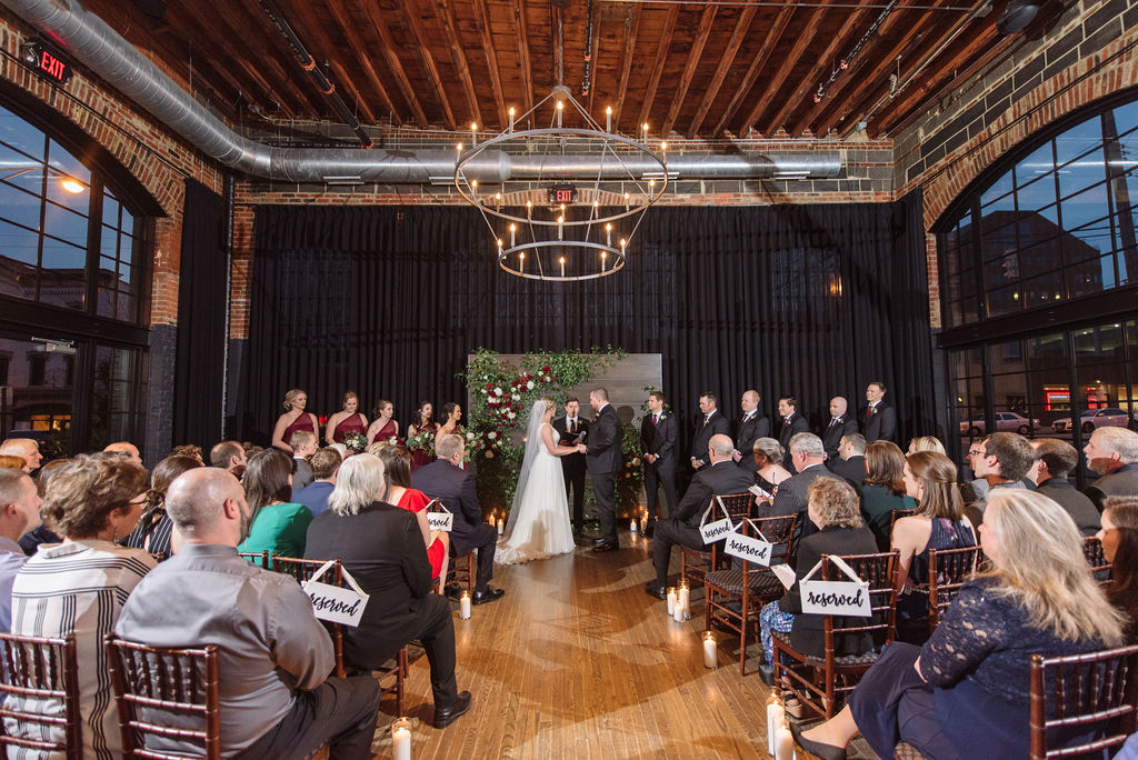 Chelsea and Chris hold hands at the front of the Social Hall at the High Line Car House during their wedding ceremony. To the left and right of them is their bridal party, all turned towards them as they make their vows. Wedding guests are seated under two-tiered candelabra chandeliers and watching the ceremony.