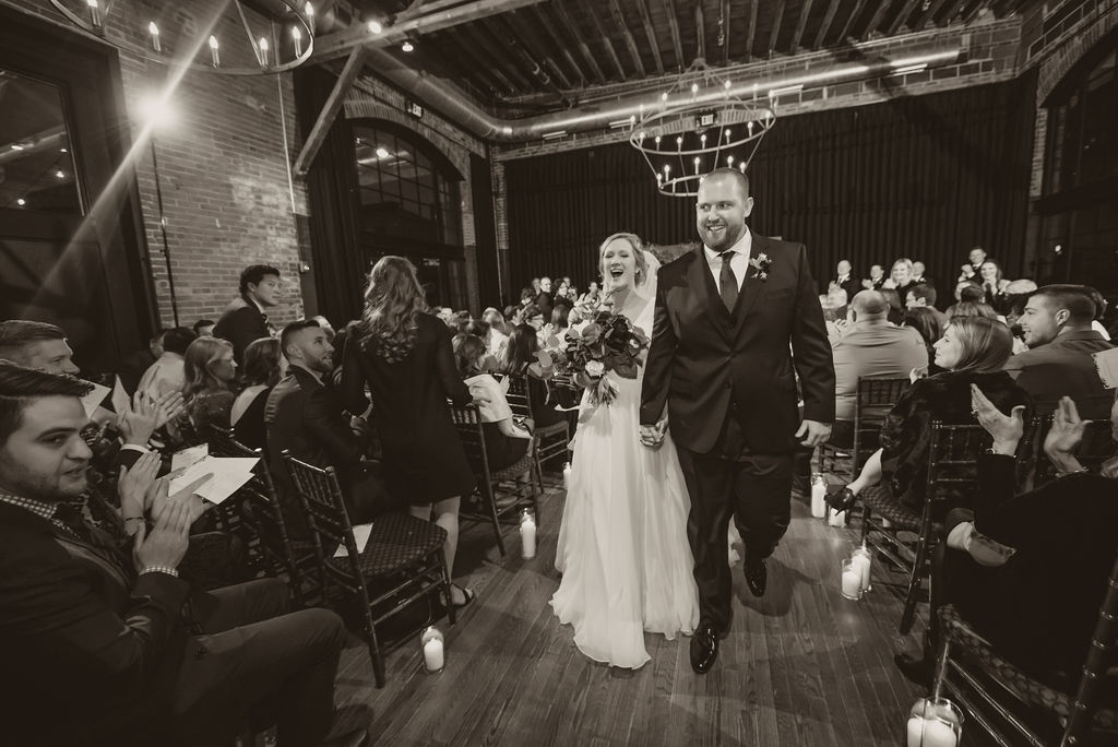 A black and white photo of a couple walking down the aisle after their ceremony in an industrial room with brick walls and two-tiered candelabra-style chandeliers. They are celebrating and smiling at their friends and family as they walk. Guests stand and applaud around them.
