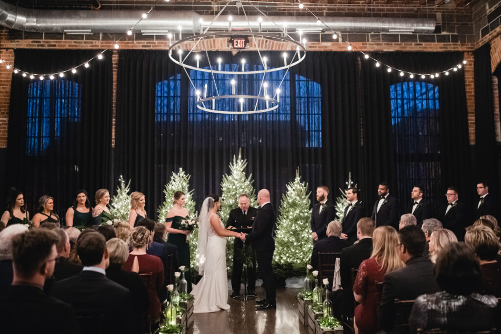 A view of the Social Hall at the High Line Car House decorated with string lights and lit evergreen trees. A couple stands at the front of the room, holding hands at their altar, as the bridal party and wedding guests watch.
