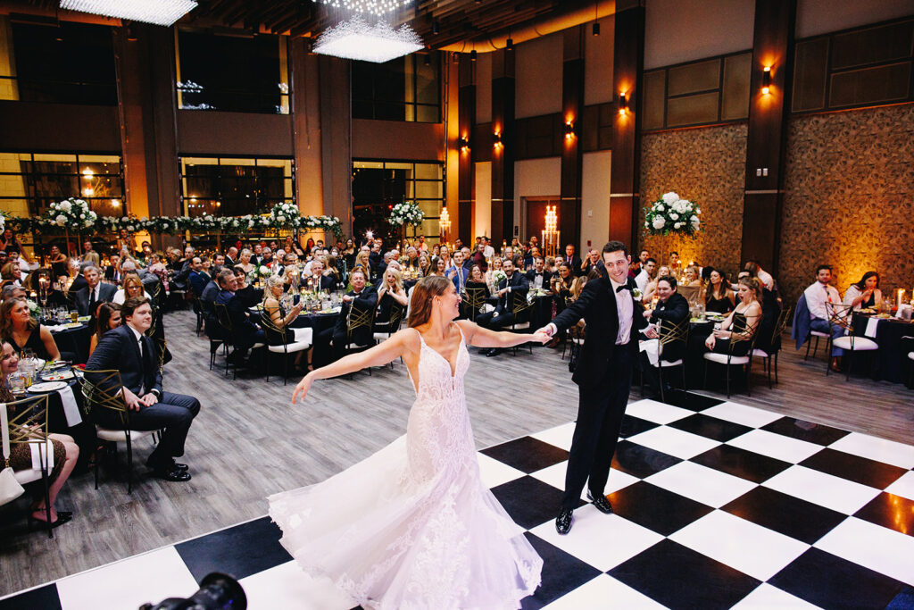 Amanda and Jeremy take over a black and white checkered dance floor for their first dance. Jeremy holds Amanda's hand as she spins out away from him, her dress spiraling out under her. Guests watch with admiration behind them in the romantically lit Social Hall at the Fives.