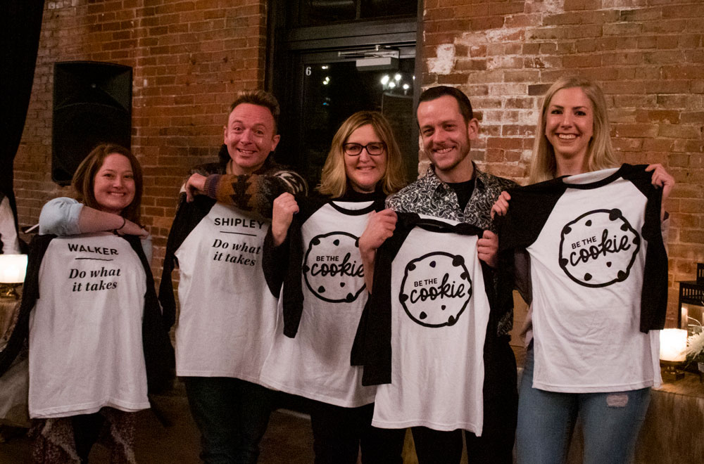 Five smiling employees are holding up henley style long sleeve tshirts. The three employees on the right are showing the front of the tshirts, which say "be the cookie". The two employees on the left are showing the backs of the shirts that say "do what it takes", with their last names above it.