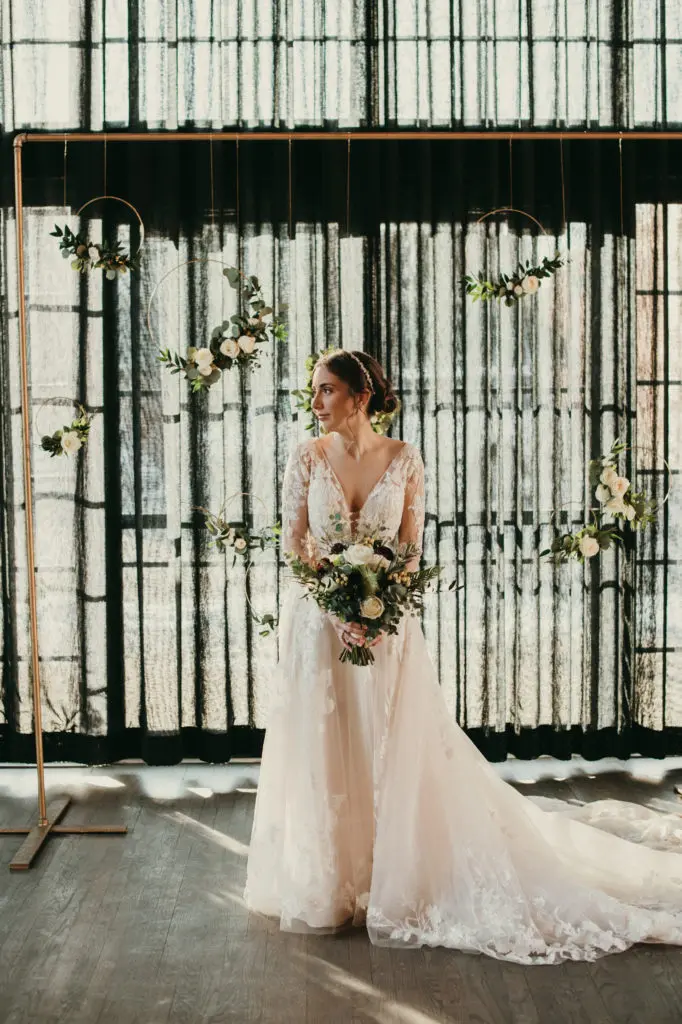 Rachel stands in front of the altar by herself, looking off to the right, holding her bouquet low. Behind her are tall dark curtains that let the sunlight shine into the room.