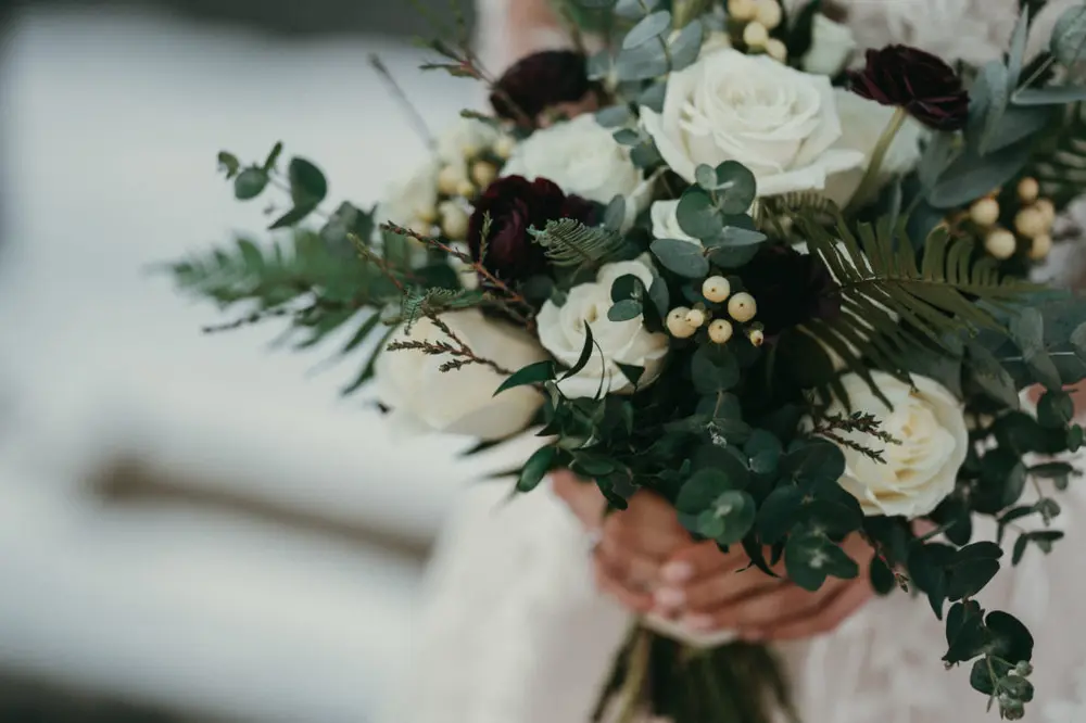 A closeup of Rachel's bouquet in her hands. The bouquet is filled with white and red roses, cushioned with frosty green accents. In the blurred background, there is snow on the ground.