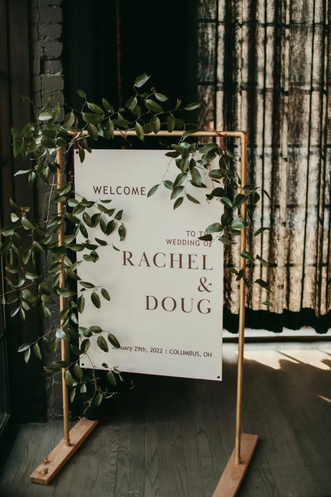 A sign at the entrance of the High Line Car House that reads "Welcome to the Wedding of Rachel & Doug". It is framed by copper pipe and accented with greenery.