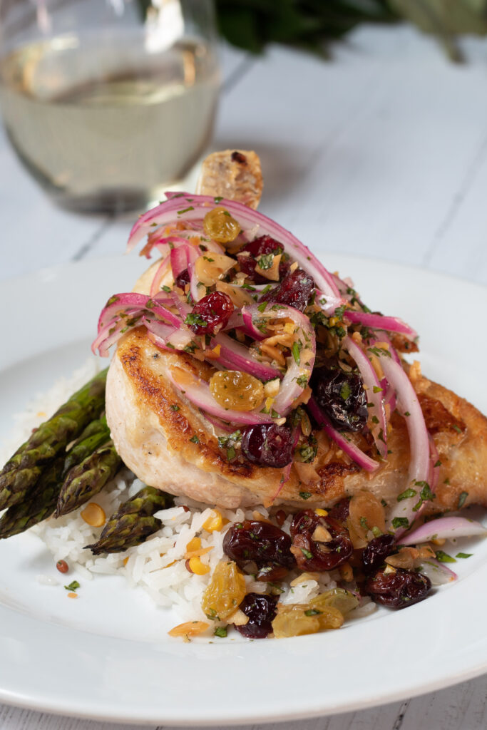 A winter menu meal from year's past sits on a white dish. It is a roasted chicken leg quarter garnished with cranberries and onions atop a bed of roasted asparagus and creamy mashed potatoes. In the background is a glass of white wine.
