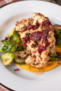 Herb Roasted Cauliflower Steak with Cranberry Sauce is on a white plate on a bed of brussels sprouts and puréed sweet potatoes.