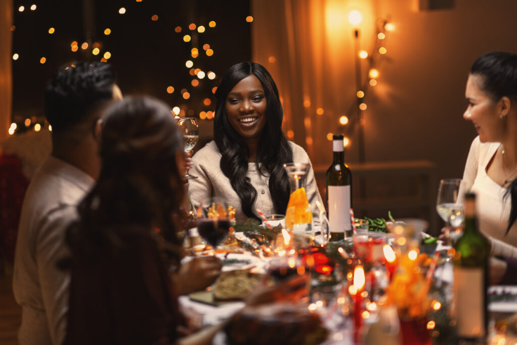 A group of friends sit around a holiday dinner table. The person facing the camera is smiling and looking at another friend. In the background are glowing stringed lights. There is a bottle of wine and wine glasses on the table.