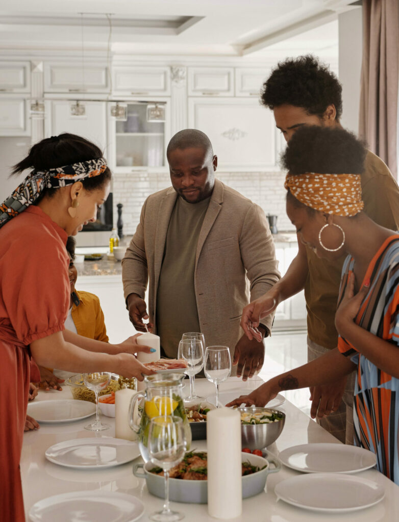 A group of family and friends gather around a white kitchen counter filled with food and plates. They are each reaching for an item on the counter and smiling.
