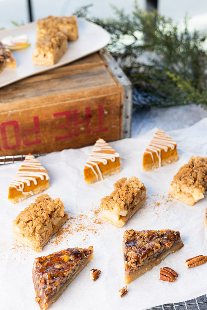 Three rows of Mini Apple, Pecan, and Pumpkin Bars sit on parchment paper on a cookie rack. Garnish of pecans and pumpkin spice is scattered throughout the parchment. In the background are assorted Mini Bars on a long white plate sitting on a wooden box and some evergreen decoration.