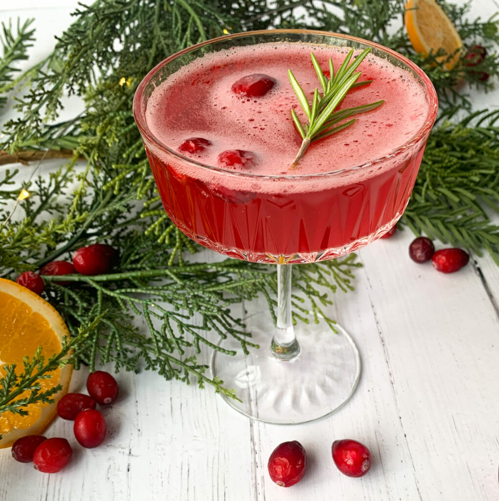A Cranberry Sparkler beverage in a coupe glass, with cranberries and a sprig of rosemary for garnish. In the background is greenery and scattered cranberries and orange slices.
