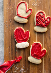 Overhead view of red Mitten Sugar Cookies in the middle of being decorated on a wooden cutting board. The two mittens towards the bottom of the frame are fully decorated, and the two cookies in the upper part of the frame are partially decorated. A bag of red icing sits off to the side, partially draped on the cutting board.