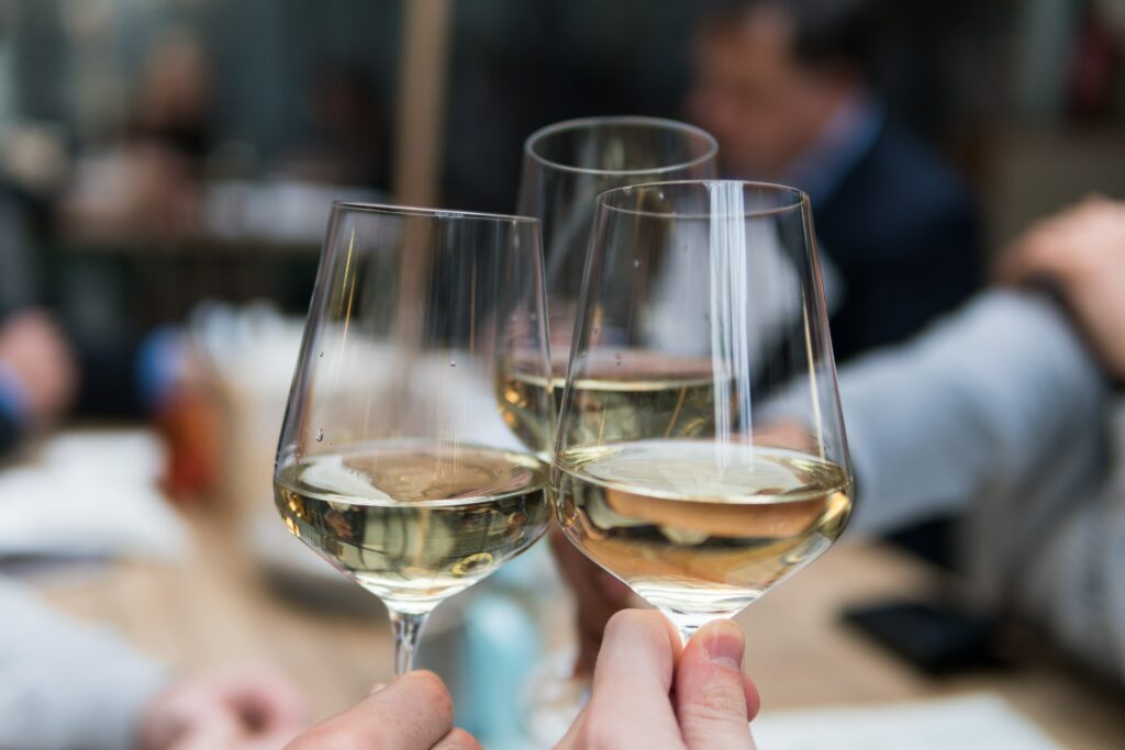 Three stemless wine glasses filled with white wine join together in a "cheers". In the background is an out-of-focus group of people around a table sharing a meal.