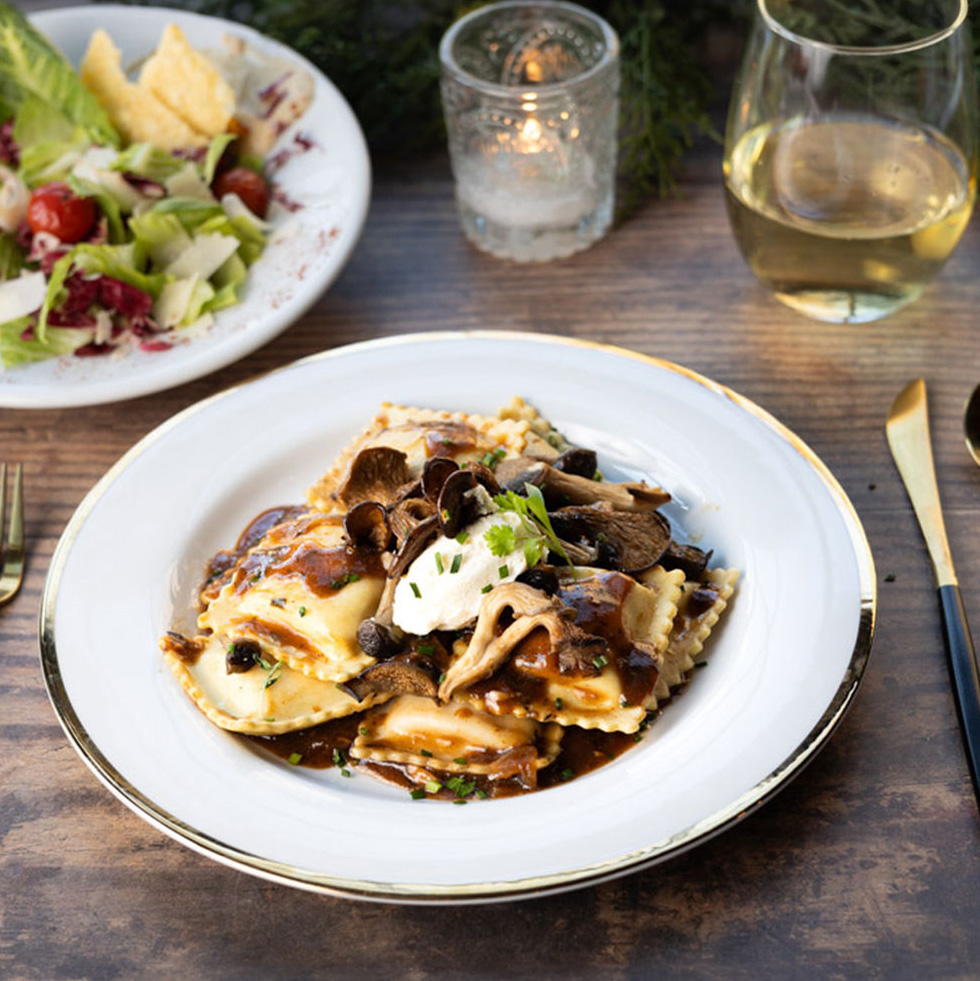 Our Wild Mushroom Ravioli sits on a white plate with gold trim. On the upper left corner of the photo is our Red Caesar Salad. Directly behind the plate is a glass of white wine and a tea light candle in a glass candle jar.