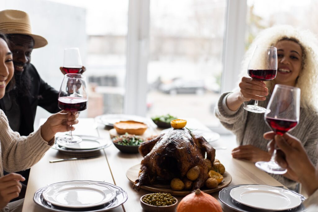 A group of friends sit at a Thanksgiving table together, their wine glasses raised in a toast. There is a turkey in the middle of the table with sides around it. Everyone is smiling and looking at each other