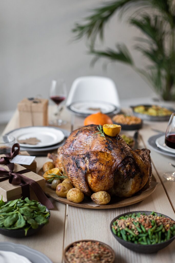 A Thanksgiving turkey sits on a platter in the middle of a table. There are sides all around: green beans, spinach, among others. A couple wine glasses are out of focus in the periphery. Two small brown boxes with brown ribbon are to the left of the turkey.
