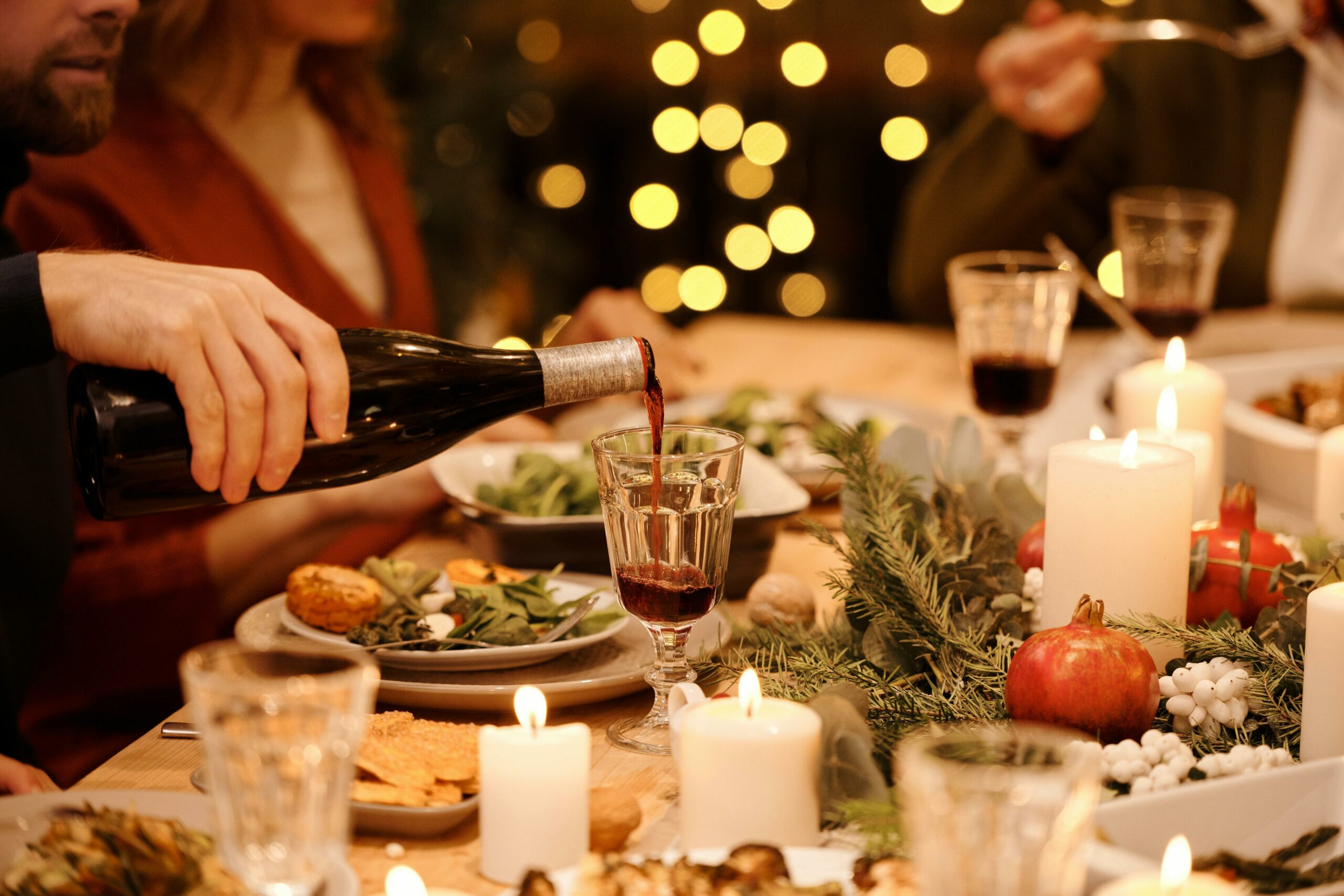 Someone out of frame is pouring a bottle of red wine into a wine glass at a holiday dinner table. There are short, white candles scattered around the table, along with table decorations such as evergreen trimmings and pomegranates. In the background are other guests at the table and out-of-focus twinkle lights.