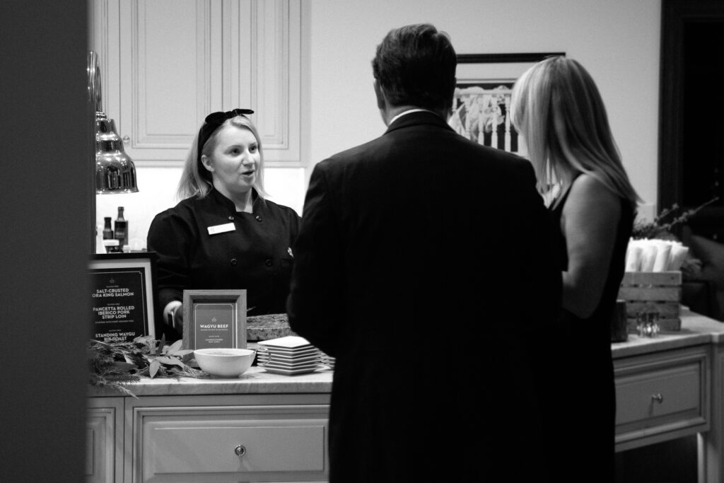 A Together & Company chef speaks with a couple of guests at a holiday house party. Chef stands behind a counter, facing the camera, and we see the guest's backs. The guests are dressed in a dark dress coat and black dress, as they look at Chef and listen.