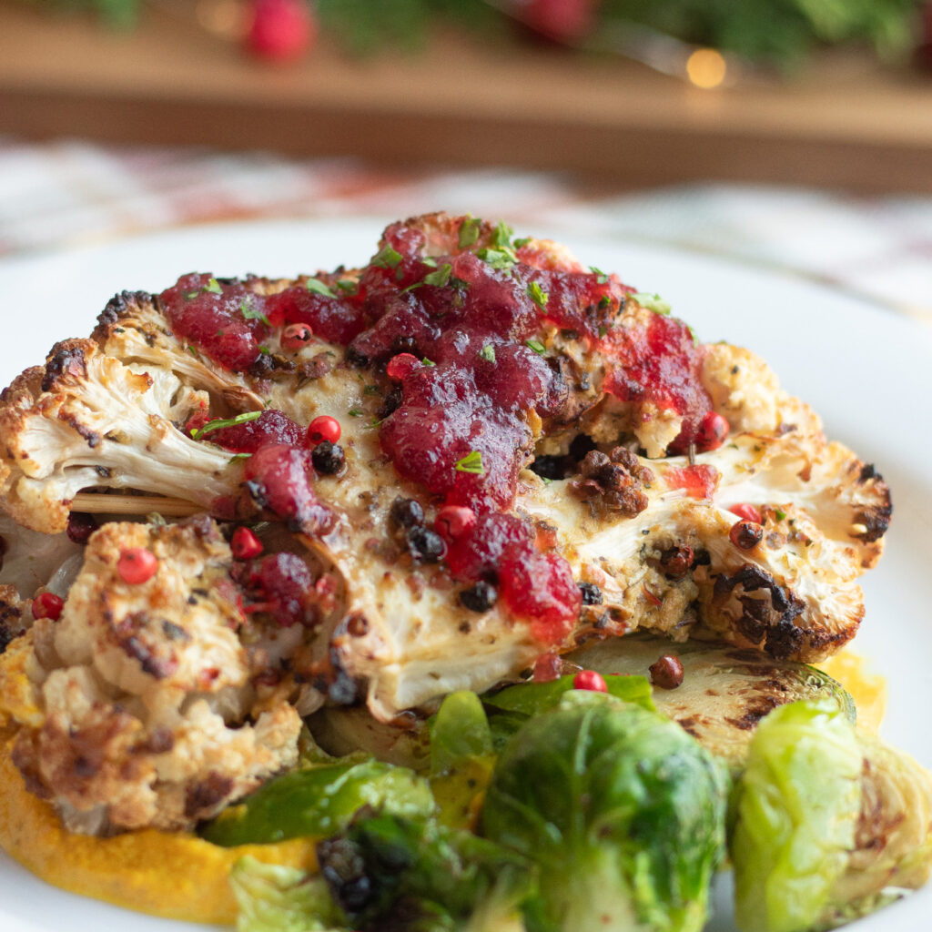 Our Herb Roasted Cauliflower Steak with a Cranberry-Orange Relish sits on a white plate next to some brussel sprouts. Under the plate is a red and white plaid tablecloth and in the background is out of focus greenery and red bauble ornaments.