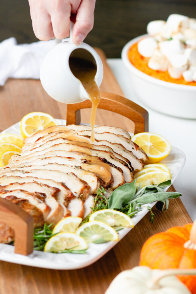 Roasted & Sliced Turkey Breast is served on a white platter with wooden handles atop a sage, rosemary, and lemon slice garnish. Turkey Pan Gravy is being poured overtop and a sweet potato dish sits in the background.