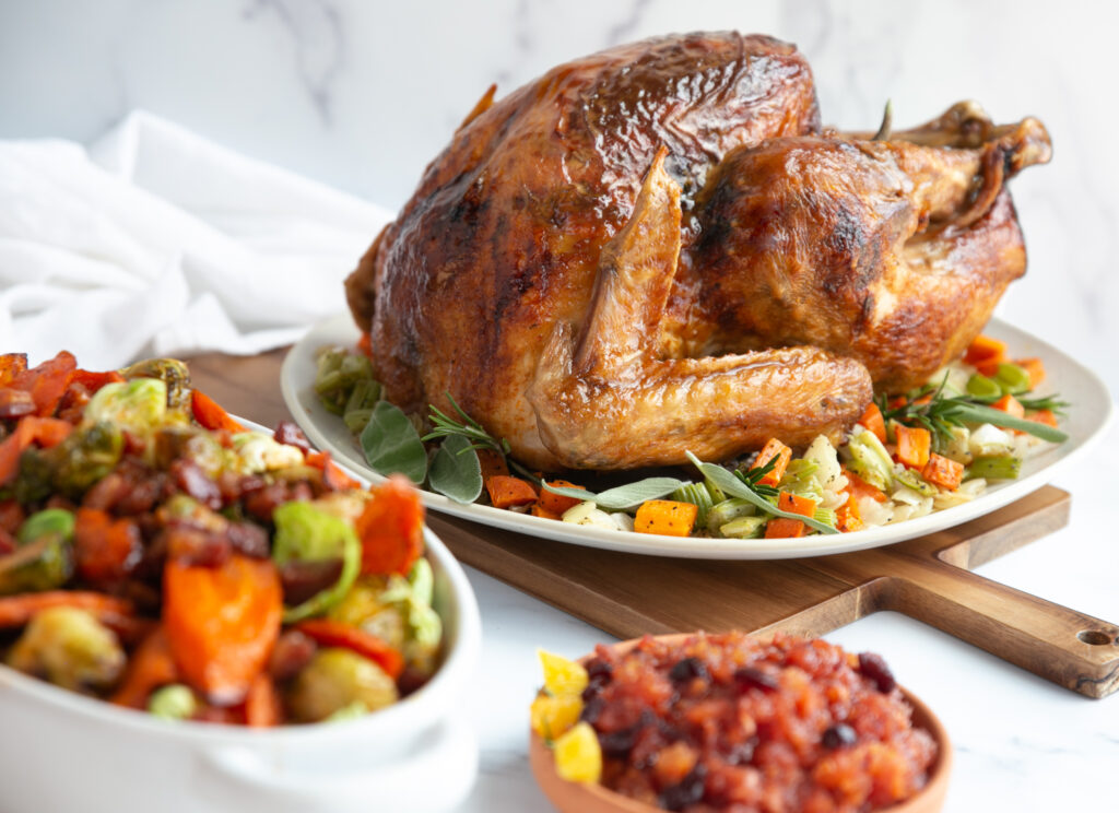 A roasted turkey sits atop a bed of vegetables on a platter. In the foreground are two other side dishes: Cranberry-Orange Relish and a root veggie mix in a casserole dish.