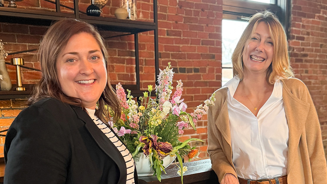 Angela and Carly stand in front of a bar setup at High Line Car House. They face each other but are turned towards the camera. There is a colorful flower arrangement between them on the bar.