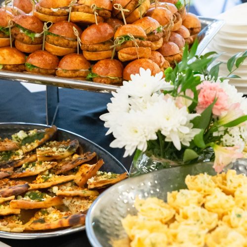 Three trays of appetizers are layered on a display table at a graduation party. The platter in the background has Together & Company Falafel Sliders, and in the foreground are platters of Seasonal Flatbreads and Mac and Cheese cups.