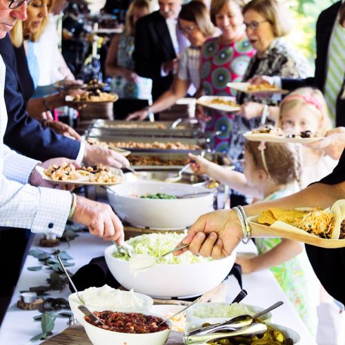 A group of guests line up on either side of a taco bar buffet at a graduation party under a tent outside in the summertime